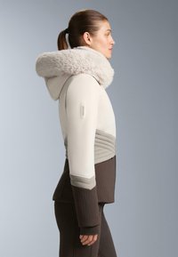 Hellfarbige Jacke mit Kapuze aus Kunstfell, mit gerippten Strukturen und farblich abgesetztem Design in Beige und Braun. Recco-Logo am Ärmel.