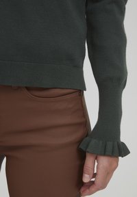 Tamnozeleni pleteni pulover s rebrastim rubom i proširenim rukavima, uparen s smešnim uskim hlačama, naglašavajući glatku teksturu i elegantan dizajn.