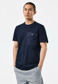 Navy blue cotton t-shirt with a chest pocket featuring a rectangular label. Short sleeves and classic round neckline. Smooth texture.