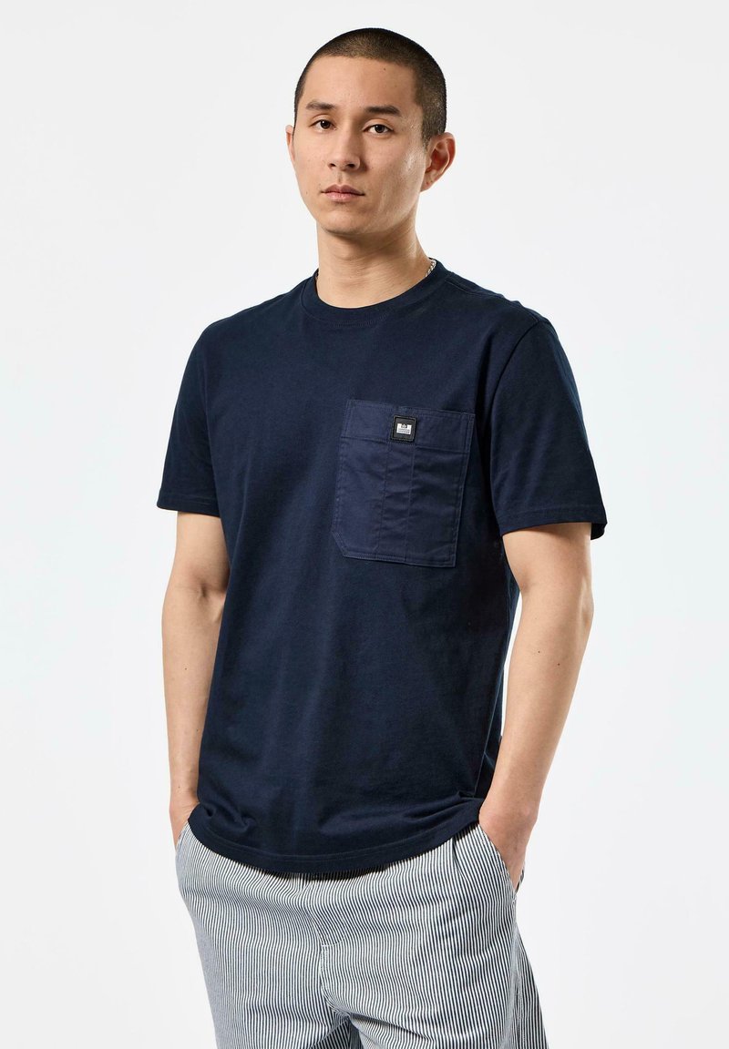 Navy blue cotton t-shirt with a chest pocket featuring a rectangular label. Short sleeves and classic round neckline. Smooth texture.