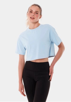 Młoda kobieta w jasnoniebieskim krótkim T-shircie i czarnych, dopasowanych leginsach z wysokim stanem, stoi na jednolitym jasnym tle.