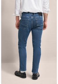 Slim-fit, ciemnoniebieskie dżinsy z lekko przetartym wyglądem, pięć kieszeni oraz widoczna metka marki na tylnej stronie pasa. Czarne buty z skarpetkami.