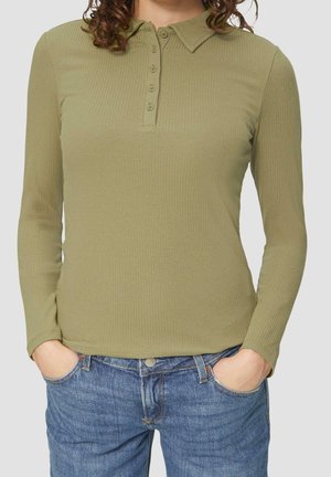 Mujer con camisa tipo polo de manga larga acanalada color verde oliva, metida ligeramente dentro de un pantalón vaquero azul, con las manos en los bolsillos.