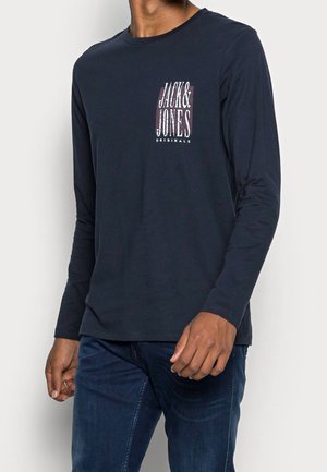 Mężczyzna w ciemnogranatowej koszuli z długim rękawem z napisem "Jack & Jones Originals" na lewej piersi, w połączeniu z niebieskimi dżinsami.