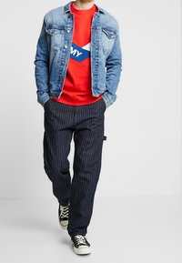 Person med en blå jeansjacka över en röd sweatshirt, randiga mörka byxor och svart-vita sneakers, går med händerna i fickorna.