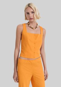 Oranje cropped tanktop met een vierkante halslijn, vijf knopen aan de voorkant en een soepele textuur. Gecombineerd met bijpassende broek en een kralenketting.