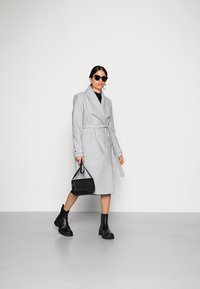 Cappotto avvolgente grigio in lana con vita aderente, maniche lunghe e rever ampi; accessorato con una borsa nera e stivaletti.
