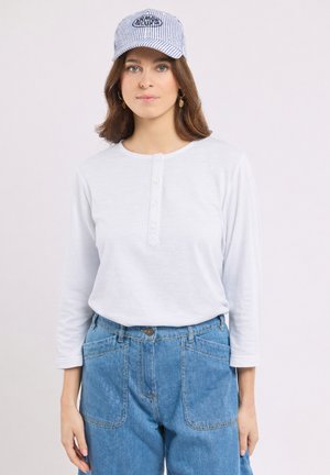 Jeune femme aux cheveux bruns mi-longs portant un t-shirt henley blanc à manches longues, un jean bleu, une casquette rayée et des boucles d'oreilles dorées.