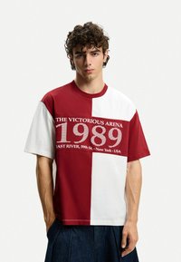T-shirt color block rosso e bianco con maniche corte, caratterizzata da un design grafico con il testo "L'ARENA VITTORIOSA 1989" sul petto.