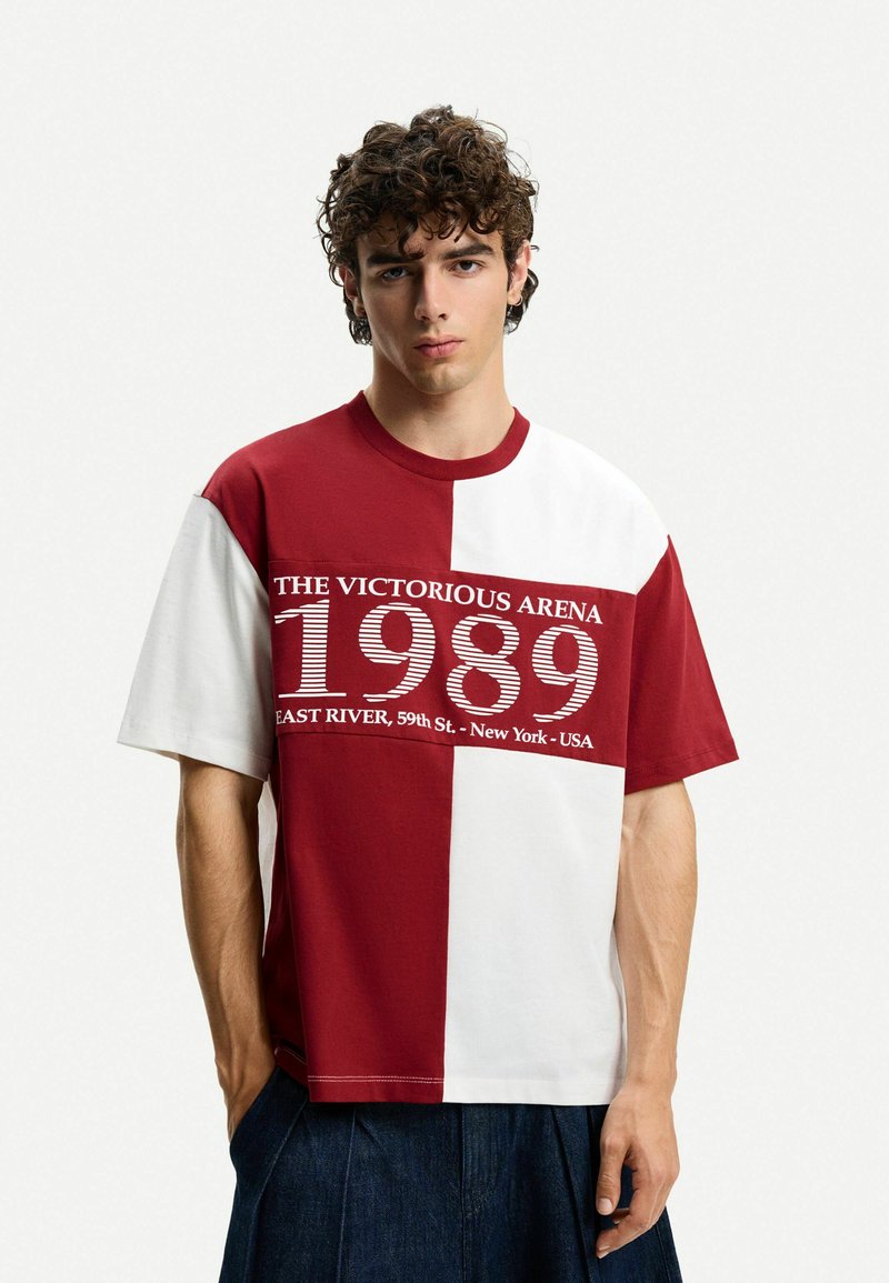 T-shirt color block rosso e bianco con maniche corte, caratterizzata da un design grafico con il testo "L'ARENA VITTORIOSA 1989" sul petto.