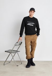 Sudadera negra con texto en blanco, pantalones cargo color beige y botas negras. Silla plegable gris cerca, situados contra un fondo neutro.