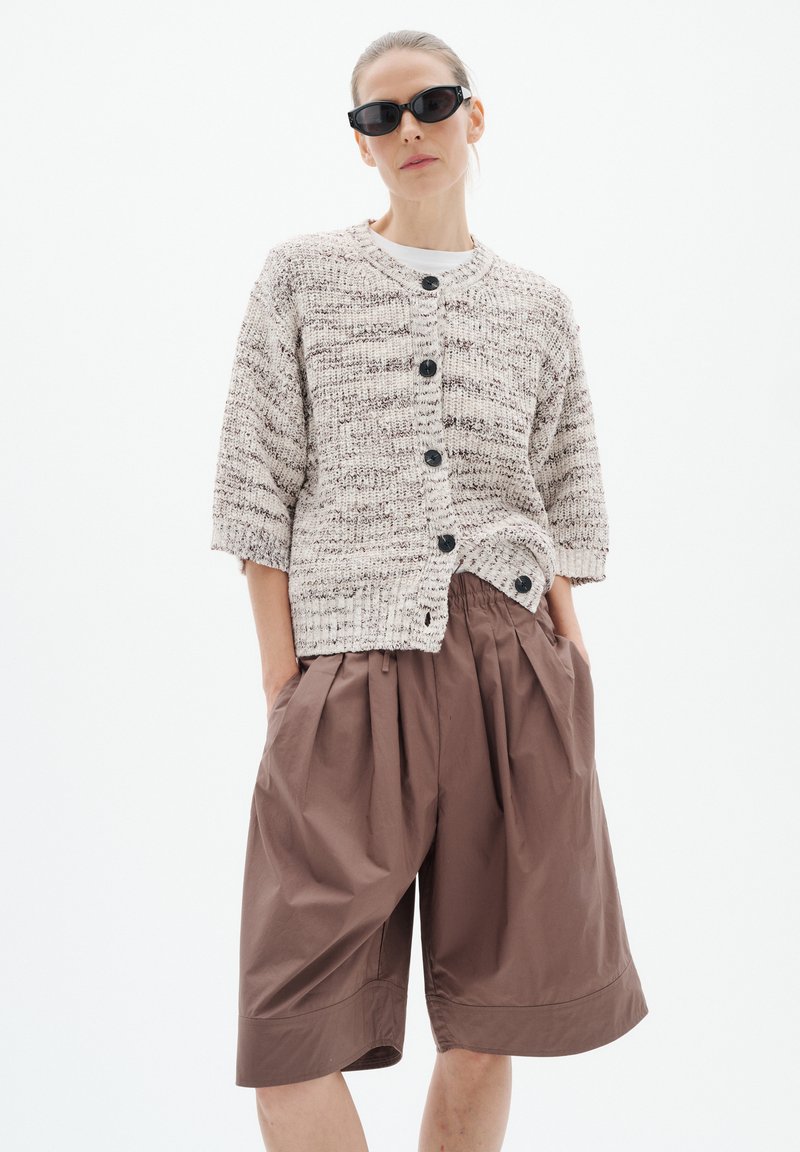 Femme portant des lunettes de soleil noires, un cardigan beige en maille boutonné et un short marron ample arrivant aux genoux, les mains dans les poches.