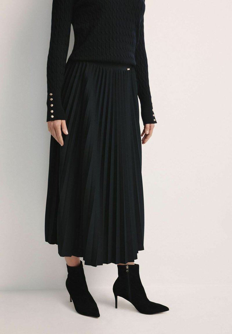 Czarna plisowana spódnica z lekkiego materiału, w połączeniu z czarnymi zamszowymi botkami za kostkę z ostrymi noskami i detalem zamka błyskawicznego z boku.