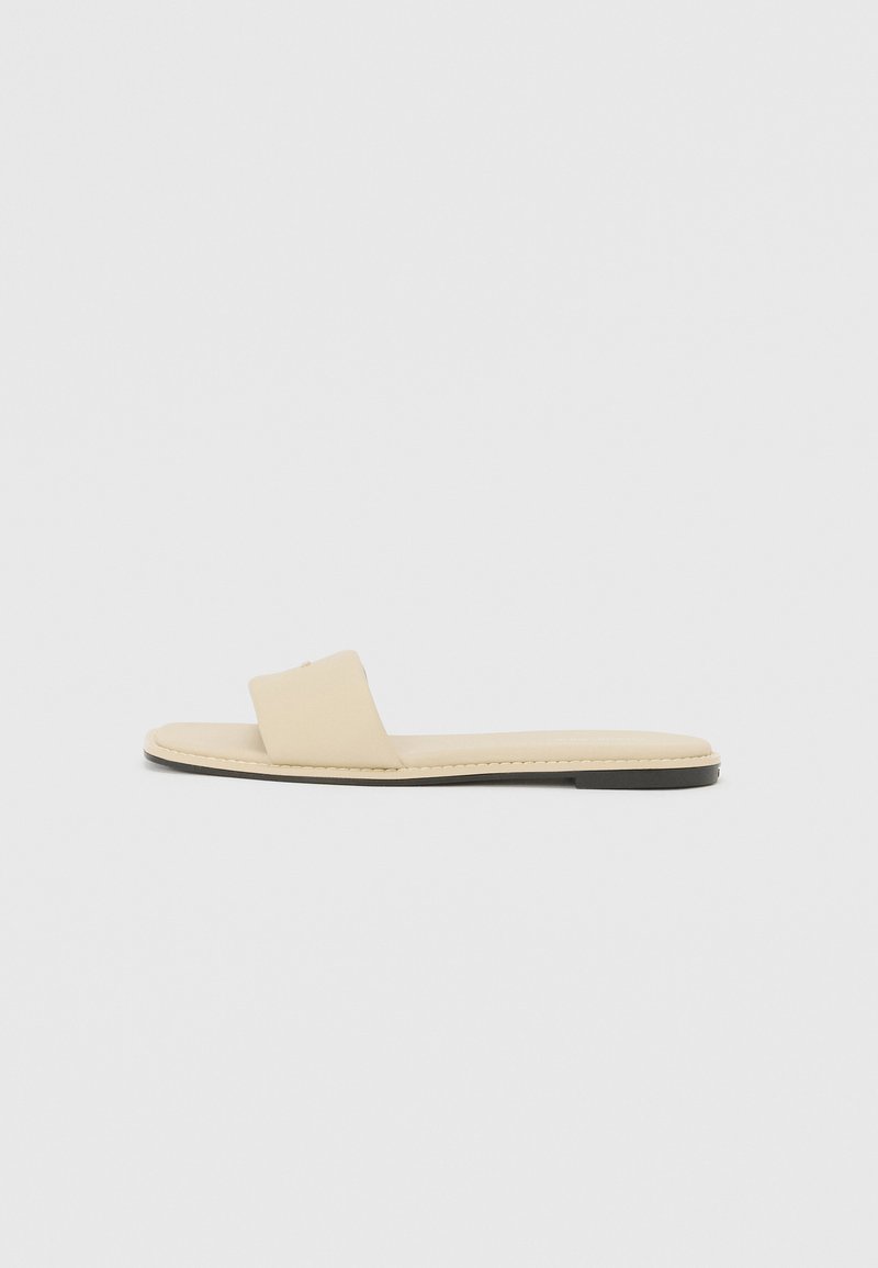 Beige flache Rutsch-Sandale mit einem breiten Einzelriemen, glattem Leder-Obermaterial, gepolstertem Fußbett und einem minimalistischen Design mit dünner Sohle.