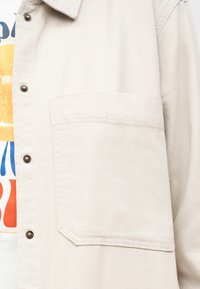 Μπεζ υφασμάτινο μπουφάν με μεγάλη μπροστινή τσέπη στο στήθος και μεταλλικά πατιλέτα κουμπιά, φορεμένο πάνω από πολύχρωμο γραφικό T-shirt.