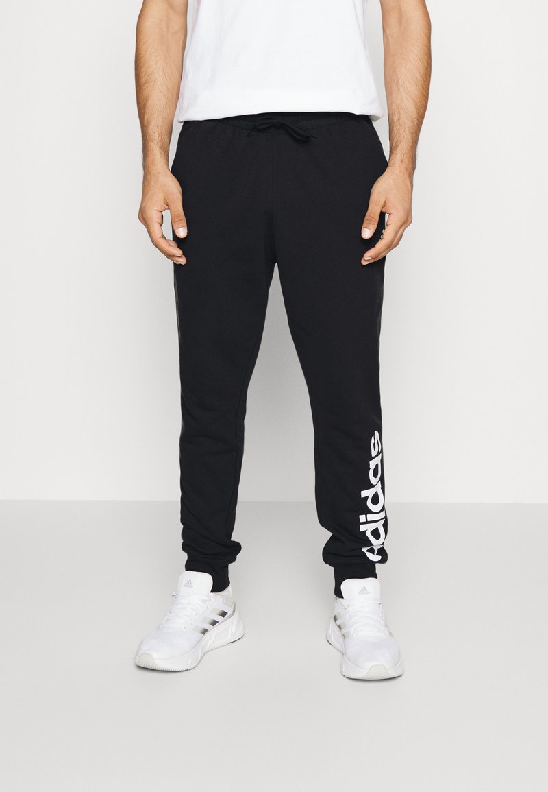 Pantalons de survêtement noirs avec un logo Adidas blanc sur la jambe gauche, dotés d'une taille élastique avec cordon et d'une coupe fuselée, portés avec des chaussures de sport blanches.