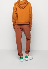 Sudadera con capucha marrón de corte casual, combinada con pantalones marrones a juego que tienen puños enrollados y zapatillas blancas con detalles en verde.