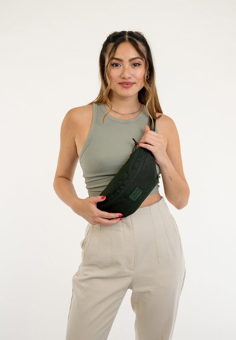 Borsa a vita di tessuto verde scuro con chiusura a zip e un'etichetta rettangolare, tenuta da una persona che indossa una canotta a coste verde chiaro e pantaloni beige.