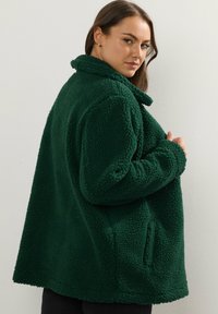Zielona polarowa kurtka o teksturowanej, pluszowej powierzchni, z dużym kołnierzem, otwartymi kieszeniami i luźnym krojem, zaprojektowana z myślą o ciepłe i komforcie.