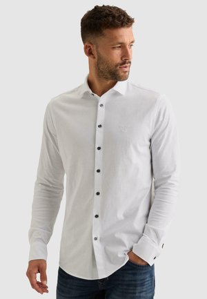 Homme aux cheveux courts et bouclés avec une barbe, portant une chemise blanche cintrée à manches longues boutonnée et un jean bleu, regardant vers la droite.