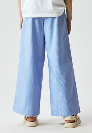 Persona, valkā gaiši zilas platas svītrainas bikses un bēšas sandales, stāv pret vienkāršu gaišu fonu.