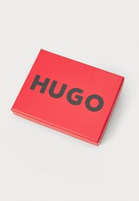 Boîte rectangulaire rouge avec une finition lisse, portant le mot "HUGO" en lettres noires en gras sur la surface supérieure.