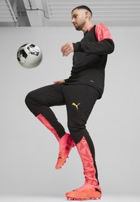 Schwarzes Sporttop mit pinkem geometrischen Ärmeln; schwarze Hose mit pinken Akzenten und gelbem Logo; pinke Fußballschuhe; schwarz-weißer Fußball.