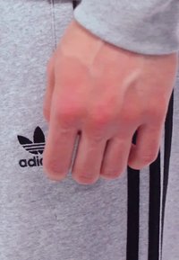 Grå sweatshirtstof viser et sort Adidas-logo med tre striber. En hånd hviler nær logoet og viser detaljer af hudens tekstur og årer.
