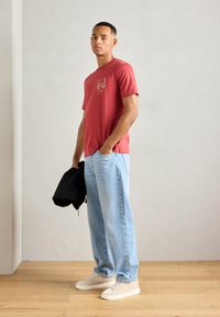 Rdeča majica s kratkimi rokavi z grafiko na prsih, v kombinaciji z svetlo modrimi širokimi kavbojkami in bež supergami. Model drži črn jopič.