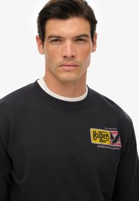 Sudadera negra con cuello redondo y puños de canalé; presenta un logotipo gráfico colorido en amarillo, rojo y negro en el pecho.