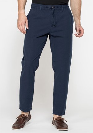 Uomo che indossa pantaloni casual blu navy con coulisse e scarpe da barca in pelle marrone con lacci, in piedi su sfondo bianco.