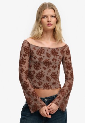 Kobieta o blond włosach ubrana w przezroczystą, brązową, kwiecistą bluzkę z długim rękawem i odkrytymi ramionami oraz ciemne jeansy, ze splecionymi rękami przed sobą.