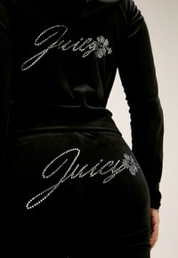 Crni baršunasti trenirka, s rhinestone "Juicy" logotipom na leđima. Uski kroj s dugim rukavima i mekanom teksturom.