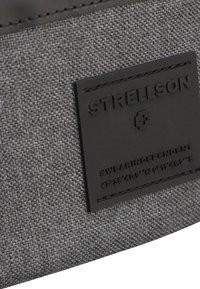 Tejido gris texturizado con una etiqueta de goma negra que presenta "STRELLSON" y coordenadas. El diseño es funcional con detalles de costura visibles.