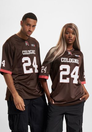 Deux personnes portant des maillots de sport Cologne marron assortis avec le numéro 24 et un pantalon noir sont debout devant un fond blanc uni.