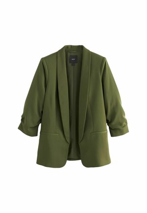 Femme portant un blazer vert olive, un débardeur noir, un jean bleu foncé, des tongs noires, et tenant une grande pochette tissée noire.