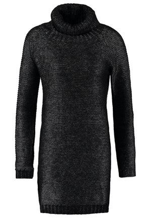 Kvinde i sort trenchcoat og sweaterkjole står med krydsede ben, holder en metallisk taske og har sorte sneakers på, foran en ensfarvet baggrund.