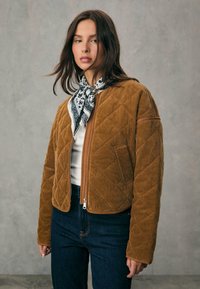 Brun kordfløyelsjacka med diamantquiltad mönster, dragkedja fram, och kontrasterande tan accenter, tillsammans med en svart-och-vit mönstrad scarf.