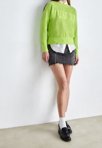 Maglione lavorato a maglia verde neon con motivi testurizzati, indossato sopra una camicia bianca con colletto, abbinato a una minigonna in denim nera e scarpe nere.