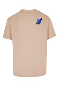 Beige bomulds t-shirt med et blåt sommerfuglegrafik trykt på den øverste ryg, korte ærmer og rund halsudskæring.