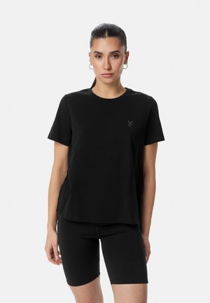 Žena s tmavými vlasmi zviazanými dozadu, oblečená v čiernom tričku s krátkym rukávom a čiernych kraťasoch nad kolená, stojaca na jednoduchom pozadí.