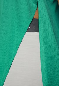 Le tissu en maille verte se divise pour révéler une partie d'une ceinture sombre sur des vêtements blancs en dessous.