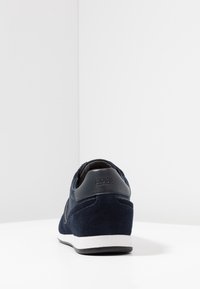 Marinblå mockaskor med ett texturerat tygpanel, vit mellansula och svarta läderdetaljer vid hälen. Har prägling av logotypdetalj.