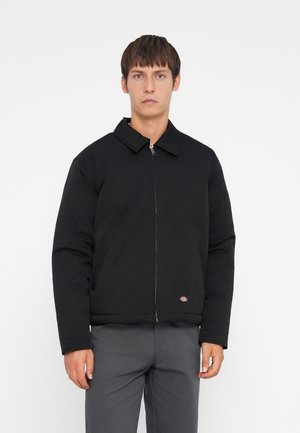 Black zip-front jacket with a pointed collar, padded texture, two side pockets, and a brand logo on the lower front hem.