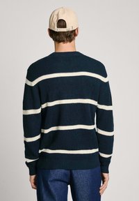 Maglioncino blu scuro lavorato a maglia con ampie righe orizzontali bianche, colletto girocollo e polsini a coste, abbinato a pantaloni in denim blu e un cappellino beige.