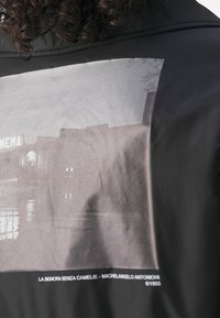 Černá bunda s velkou černobílou fotografií na zádech, zobrazující filmovou scénu s textem pod ní. Hladký, lehký materiál.