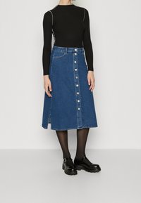 Jeanskjol med knappfram, knälång, blå färg, har sidofickor, bärs med svart långärmad topp och träskor.