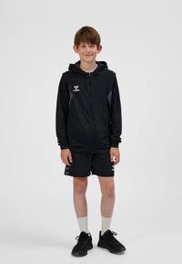 Schwarze Sportjacke mit Kapuze und Reißverschluss, mit grauen Akzenten, kombiniert mit schwarzen Shorts. Schwarze Sneaker und weiße Socken vervollständigen das Outfit.