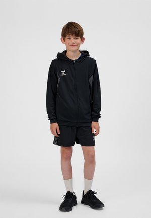Schwarze Sportjacke mit Kapuze und Reißverschluss, mit grauen Akzenten, kombiniert mit schwarzen Shorts. Schwarze Sneaker und weiße Socken vervollständigen das Outfit.