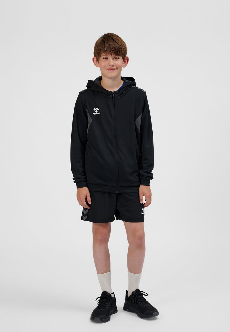 Schwarze Sportjacke mit Kapuze und Reißverschluss, mit grauen Akzenten, kombiniert mit schwarzen Shorts. Schwarze Sneaker und weiße Socken vervollständigen das Outfit.